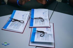 | The Calabash Newspaper Three documents bound with string and blue headers spread on a light conference table; hands are visible at the top; SLCAA logo bottom-left.