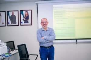 Man with glasses and a patterned shirt stands with arms crossed in a classroom, a projector screen showing an 'Exercises' slide behind him.