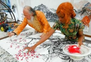 Two artists, an elderly man and a woman in bright clothing, painting a large ink-and-red blossom landscape on a floor-spread scroll.