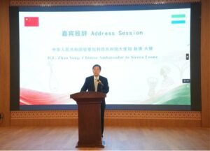 Man in a suit giving a speech at a wooden podium on a stage, with a large screen behind displaying 'Address Session' and a Chinese flag.