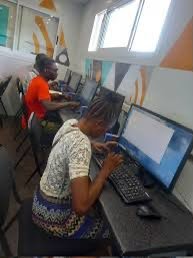 Group of students using desktop computers in a bright classroom; foreground woman in a patterned dress typing at a computer.