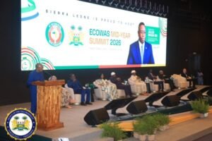 Panel discussion on stage at ECOWAS Mid-Year Summit 2026; speaker at a wooden podium with panelists seated behind, large screen branding in the background.