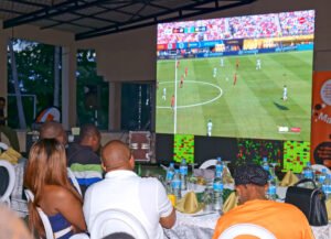 People seated at tables watching a large soccer match on a wall-mounted screen in a dining area, with bottles and place settings on the tables.