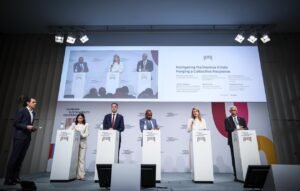 Six panelists stand at white podiums on a stage during a conference, with a large screen showing a slide about navigating the Hormuz Crisis behind them
