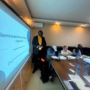 Presenter in a yellow shirt and dark suit stands beside a projection screen that reads 'Sierra Leone Railways Regulator Framework' in a conference room.