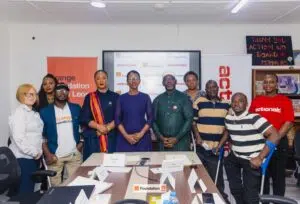 Group of diverse adults standing behind a conference table in a meeting room with Orange Foundation and actionaid branding in the background.