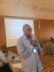 | The Calabash Newspaper Male presenter wearing glasses and a striped shirt speaks into a microphone in a conference room with a projection screen behind him.