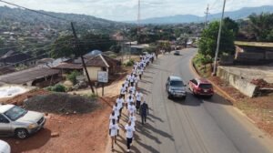 | The Calabash Newspaper Large group of people in white t‑shirts marching along a curved road through a hilly town, with cars parked and houses on either side.