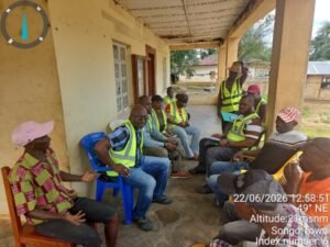 Group of men in high‑visibility vests seated on chairs along a covered porch of a yellow building, with one man standing nearby, suggesting a community meeting or briefing.