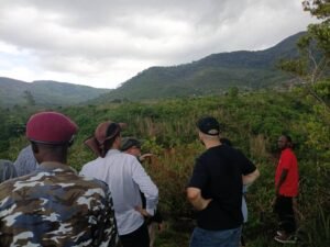 Group of people standing on a hillside, looking toward distant mountains and greenery under a cloudy sky, as if on a tour or hike.