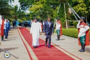 Two men in formal attire walk down a red carpet flanked by ceremonial guards in red-and-green uniforms, tropical trees behind them.