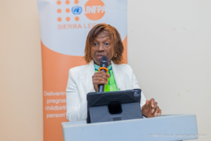 Woman in a white blazer speaks into a handheld microphone at a podium, with a UNFPA Sierra Leone banner in the background.