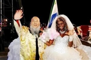 Joyful wedding moment: bride in a white gown and veil beside an elderly man in a gold ceremonial robe, both waving at night with a flag in the background.