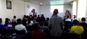 Audience seated in a small conference room watching a presentation projected on a screen at the front.