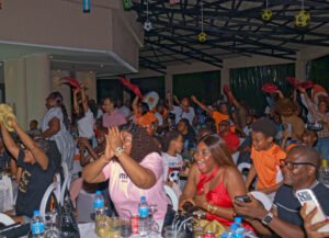 | The Calabash Newspaper Crowd of people at a lively indoor party, many raising hands and smiling, with tables of drinks in front and soccer-ball decorations overhead.