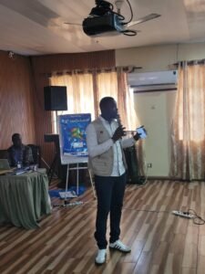 Research Officer Amadu Kamara Labor | The Calabash Newspaper A man in a light vest speaks into a microphone in a conference room, with a projector overhead and a poster board nearby.