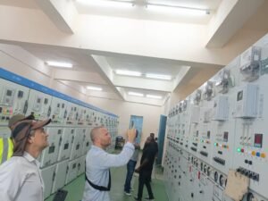 | The Calabash Newspaper Group of people touring a room full of electrical control panels with numerous switches, gauges, and indicator lights; one man photographs the panels as others look on.