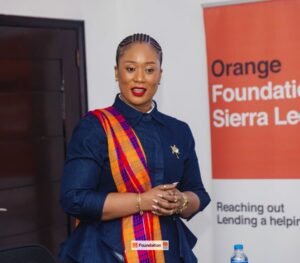 Woman in denim shirt with an orange plaid scarf speaking at an event, Orange Foundation banner visible in the background.