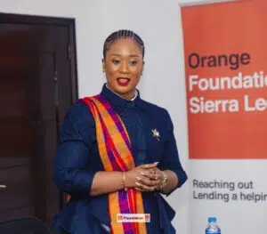 Woman in denim shirt with an orange plaid scarf speaking at an event, Orange Foundation banner visible in the background.