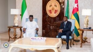 | The Calabash Newspaper Two men sit in a formal reception room with national flags behind them, a wooden emblem on the wall, and a floral centerpiece on a coffee table.