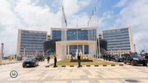 | The Calabash Newspaper Front view of the ECOWAS headquarters, a modern building with a curved glass entrance, flagpoles, cars in the foreground, and people walking around a circular garden plaza.