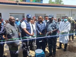 | The Calabash Newspaper Officials and community members at a ribbon-cutting ceremony outside a white building with barred windows; a uniformed officer and a woman cut the ribbon.