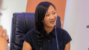 Smiling woman with a long side braid speaks into a microphone during a panel discussion, seated in a high-back chair