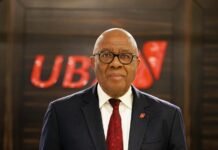 Notification of Retirement of Group Chairman of United Bank for Africa and Appointment of Successor Professional portrait of an older man in a navy suit, white shirt, and red tie, wearing glasses, standing indoors with a red UBS logo in the background