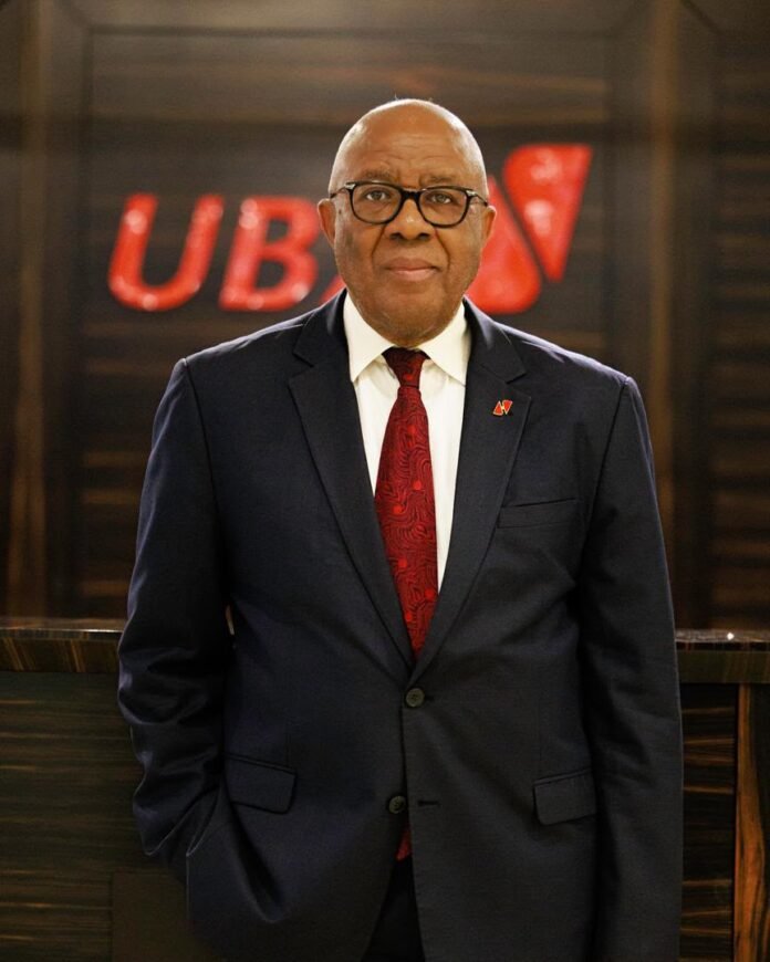 Professional portrait of an older man in a navy suit, white shirt, and red tie, wearing glasses, standing indoors with a red UBS logo in the background
