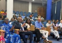 NatCA Marks 20 Years, Pledges Consumer-Focused Digital Future Audience members seated in a conference hall with blue upholstered chairs, some clapping and looking toward the stage. Front row includes a woman in a blue patterned outfit and a man in a suit.