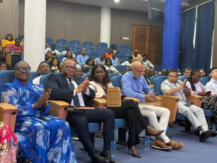 Audience members seated in a conference hall with blue upholstered chairs, some clapping and looking toward the stage. Front row includes a woman in a blue patterned outfit and a man in a suit.