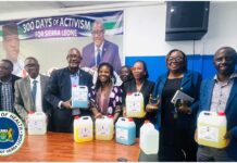 Health Ministry Begins First-Ever Local Production of Infection Prevention Supplies Group of nine people standing behind a table with large jugs of colored liquids, in a conference room with a Sierra Leone Ministry of Health banner/logo visible in the background and on the table.