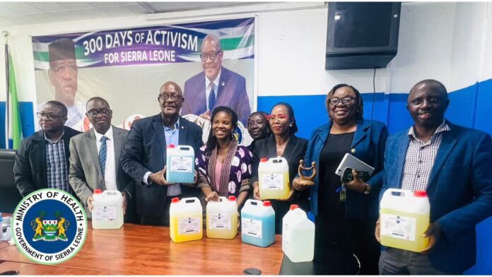 Group of nine people standing behind a table with large jugs of colored liquids, in a conference room with a Sierra Leone Ministry of Health banner/logo visible in the background and on the table.