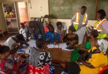 Caritas Freetown Trains Disaster Committees to Strengthen Emergency Preparedness in Vulnerable Communities Community workshop: diverse group seated around a table filling forms with volunteers in reflective vests guiding them, in a classroom with a chalkboard.