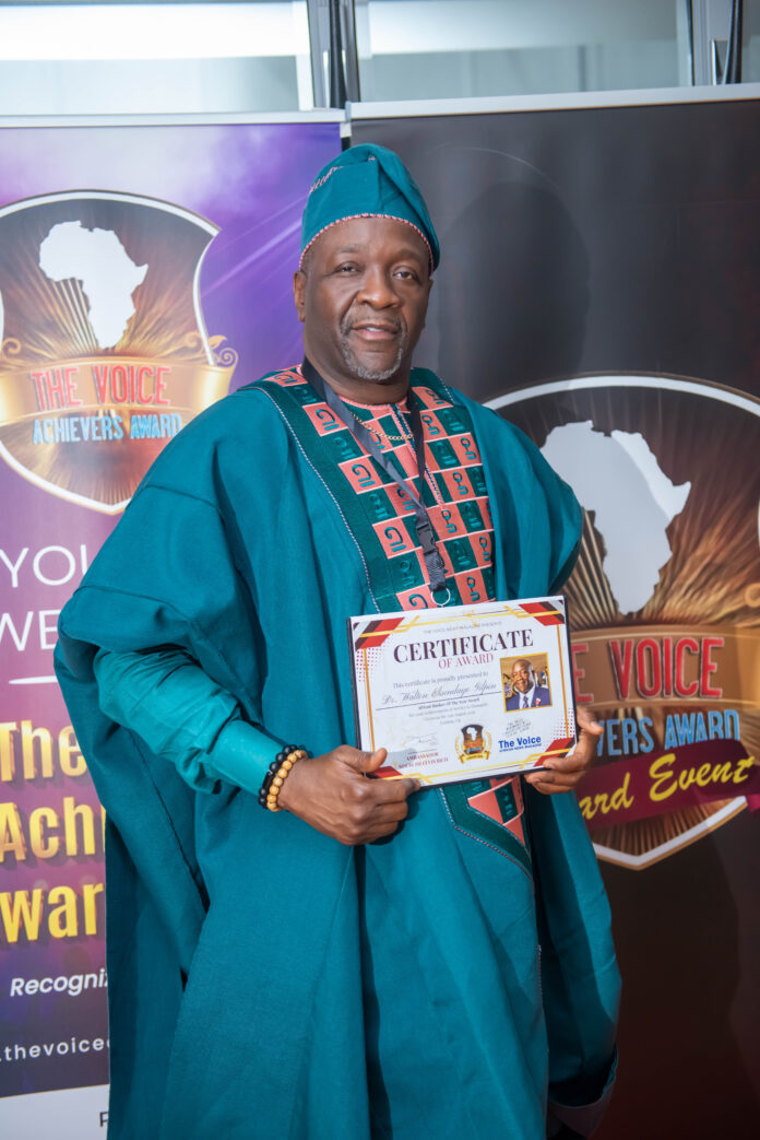 Man in teal traditional attire holding a certificate at an awards event, backdrop shows Africa map and 'The Voice Achievers Award' banner