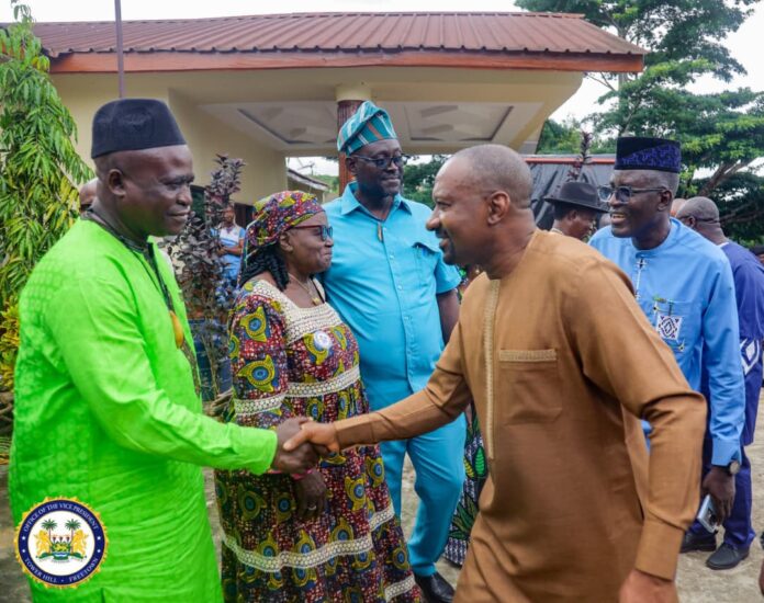 Dignitaries greet each other with a handshake outside a building, wearing bright traditional outfits and head wraps.