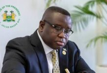 PERSPECTIVE – Why President Bio Should Wait Before Signing the Constitutional Amendment Bill A man in a dark suit signs a document at a glass desk, with an official presidential emblem in the background.