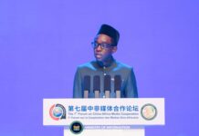 Information Minister Demands Greater African Voice in Global Media and Technology Man in traditional teal attire and a fez speaks at a podium with multiple microphones during the 7th Forum on China-Africa Media Cooperation, with a blue stage backdrop and conference banners behind him.