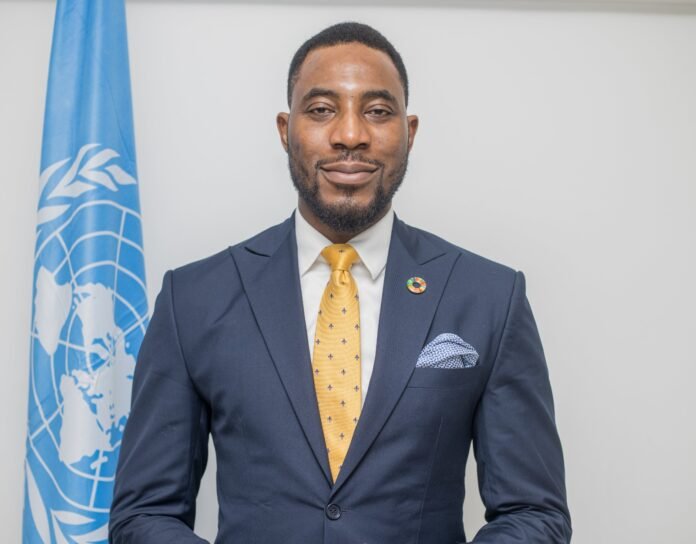 Portrait of a man in a navy suit and yellow tie, with a UN flag in the background.