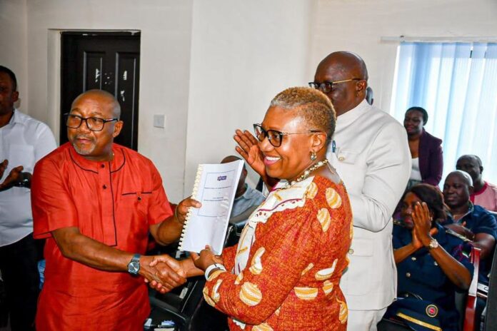 Man in red shirt shakes hands with smiling woman in orange patterned dress as she receives a document; others applaud in a bright room behind them.