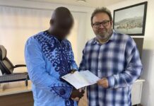 Kenema Businessman Wins NLe82,800 in Mercury International Lotto Draw Two men shake hands in an office as they exchange a document; one wears a blue traditional outfit, the other a blue plaid shirt (face blurred for one person).