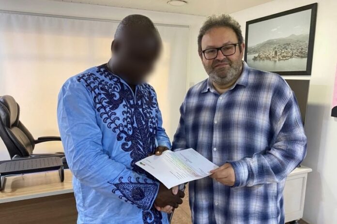 Two men shake hands in an office as they exchange a document; one wears a blue traditional outfit, the other a blue plaid shirt (face blurred for one person).