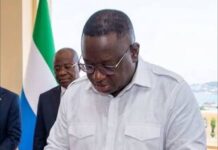 President Bio Faces Growing Calls to Withhold Assent to Constitutional Amendment Bill Man in a white shirt signs a document on a wooden table, with a flag and another man watching in the background.