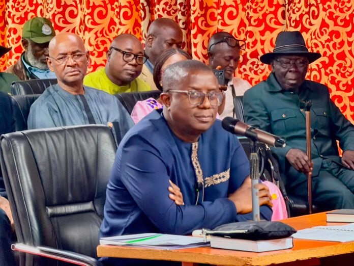 Man in blue traditional attire speaks into a microphone at a conference table, with colleagues seated behind him.
