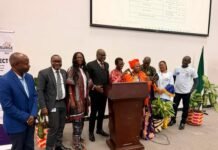 EU, Freetown City Council Launch €505,246 SURGE Project to Rebuild King Jimmy Market Group of professionals standing behind a podium at a formal event in a conference room, with a large screen overhead and banners nearby.