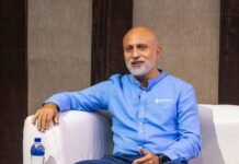 Africell CEO Shadi Gerjawi Puts Customer Satisfaction at the Heart of Service Delivery Portrait of a bald man with a white beard in a light blue shirt seated on a white sofa, interview setting with water bottle on a glass table and sponsor logos visible in the corner, afr money and Africell banners.