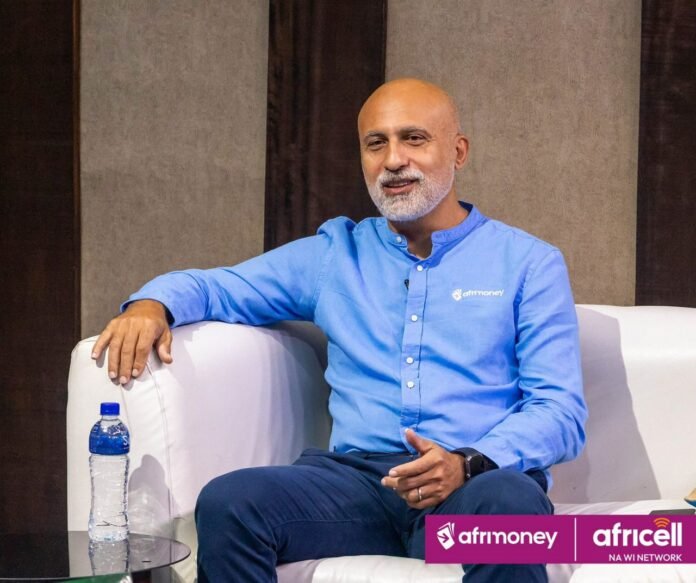 Portrait of a bald man with a white beard in a light blue shirt seated on a white sofa, interview setting with water bottle on a glass table and sponsor logos visible in the corner, afr money and Africell banners.