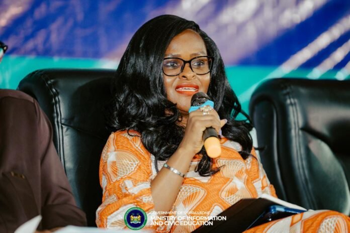 Woman in an orange patterned dress speaks into a microphone during a panel, logo of the Ministry of Information and Civic Education visible at the bottom.