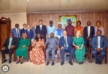 Vice President Backs Latter-Day Saints Church’s Plan to Create Hundreds of Jobs for Sierra Leonean Youths Group of formally dressed adults posed for a photo in a meeting room, front row seated and back row standing, with a framed portrait behind them and a round emblem on the floor left corner.