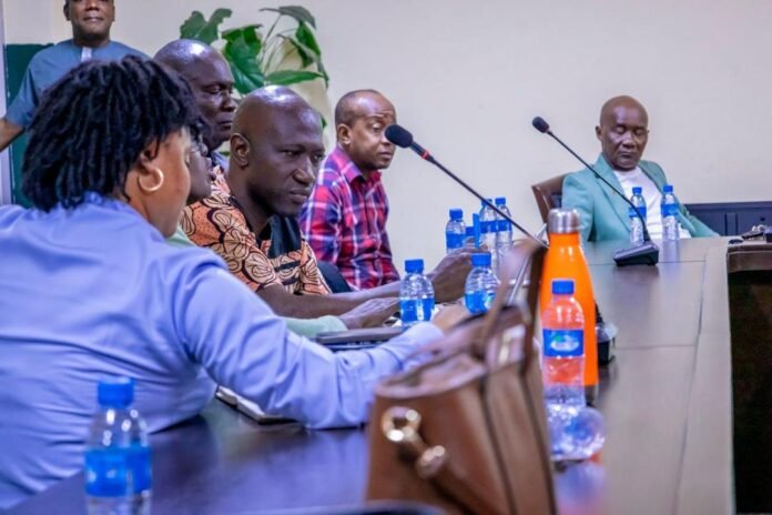 A panel of six people seated at a conference table with microphones, water bottles, and a bright orange flask in the foreground, discussing.